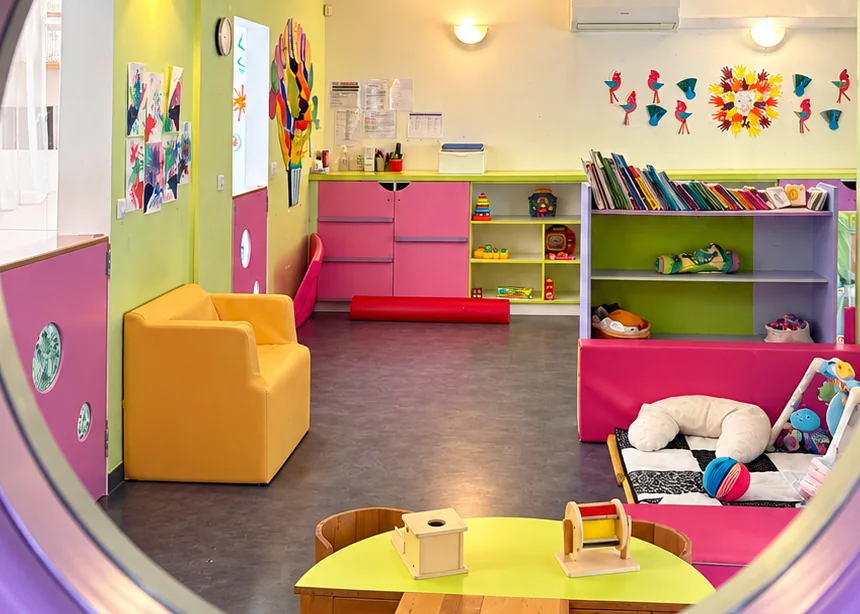 Salle de vie colorée vue par un hublot, avec fauteuil orange, bibliothèque, tapis d’éveil et jeux en bois chez Bonbon