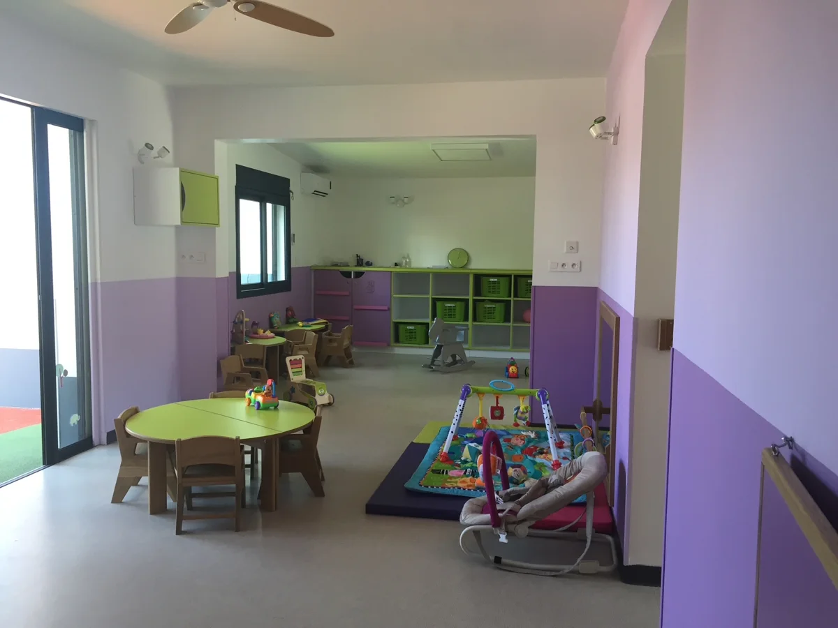 Salle de vie de la micro-crèche Caramel à Saint-Pierre, aux murs lilas, avec tables rondes vert anis, tapis d’éveil et transats