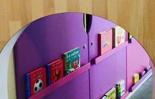 Coin lecture des Makis vu à travers un hublot, avec des albums posés face visible sur un présentoir mauve