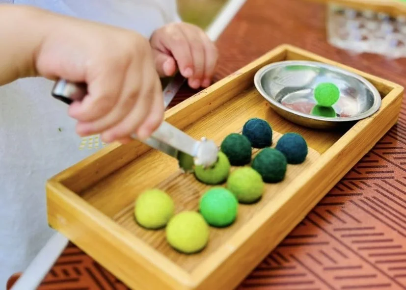 Atelier Montessori chez Pitchou : la main d’un enfant transvase des boules de feutrine avec une pince, sur un plateau en bois